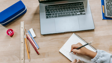 Een bureau met laptop erop, een persoon maakt een notitie.
