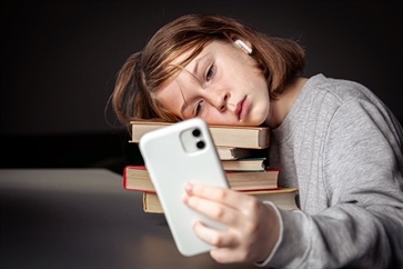 Leerling ligt met hoofd op stapelboeken en kijkt op smartphone.