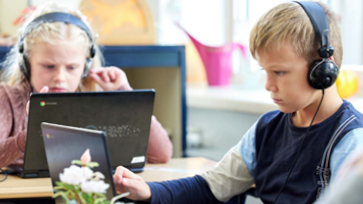 twee basisschoolleerlingen zitten met koptelefoon in de klas en kijken naar hun laptop.