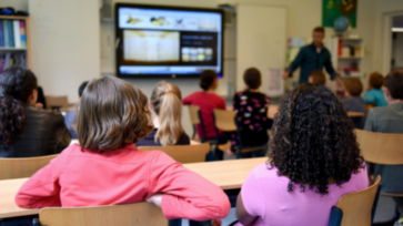 Leerlingen zitten in de klas, de leraar geeft uitleg naast het digibord.