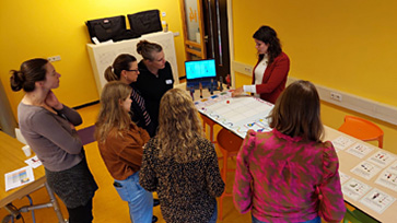 Groep mensen staat rond een tafel met kaarten en een scherm in een ruimte met gele muren.