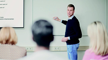 Leraar staat voor een whiteboard, en geeft uitleg aan een groep volwassenen.